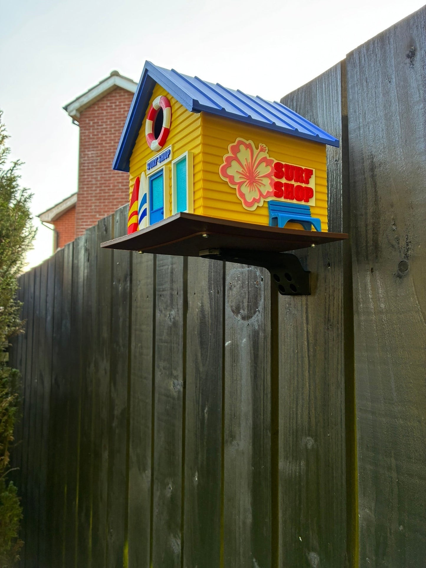 TL3D Prints yellow 3D printed surf shop birdhouse with blue roof, surfboard, and hibiscus flower detail.