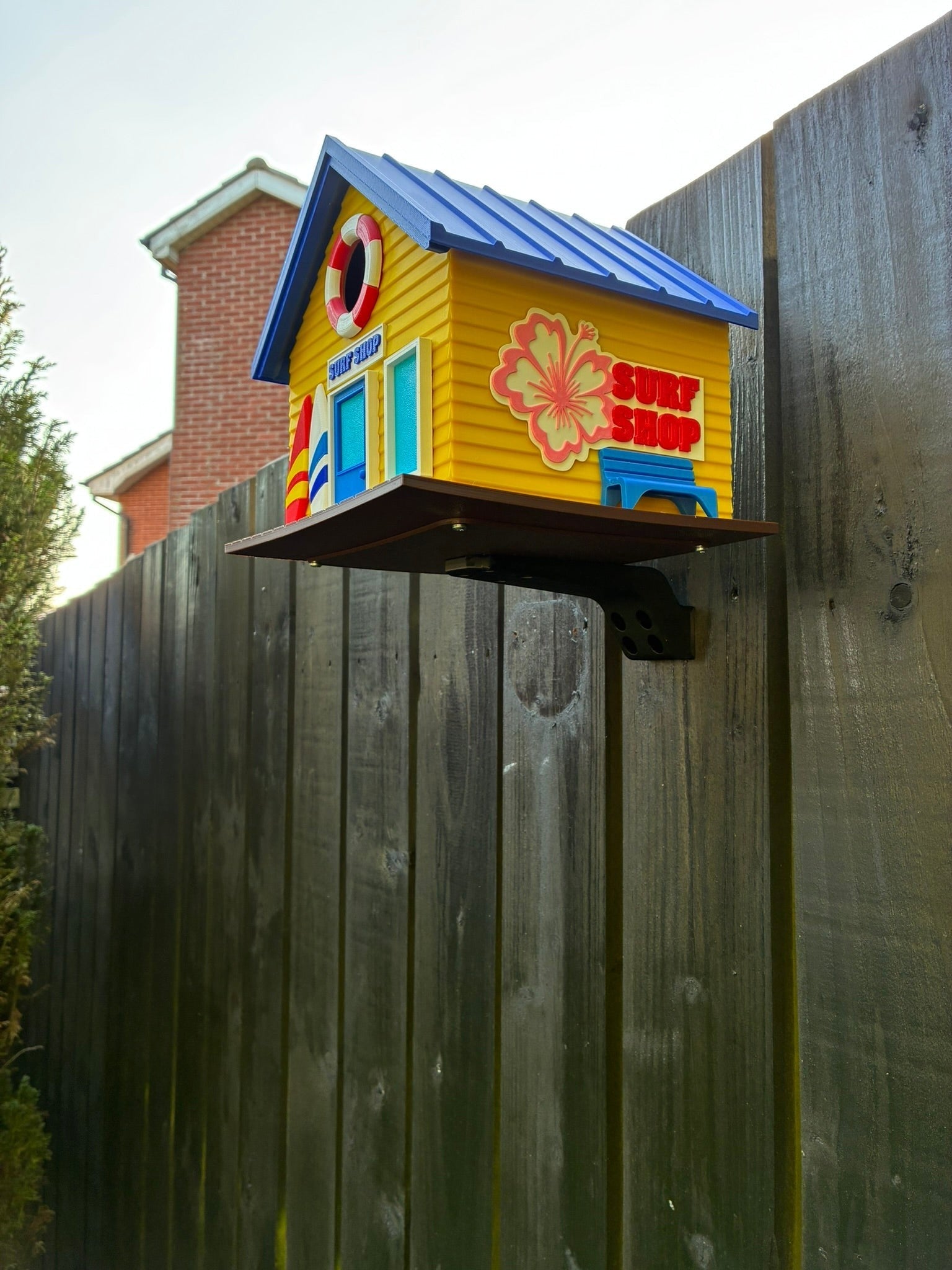 TL3D Prints yellow 3D printed surf shop birdhouse with blue roof, surfboard, and hibiscus flower detail.