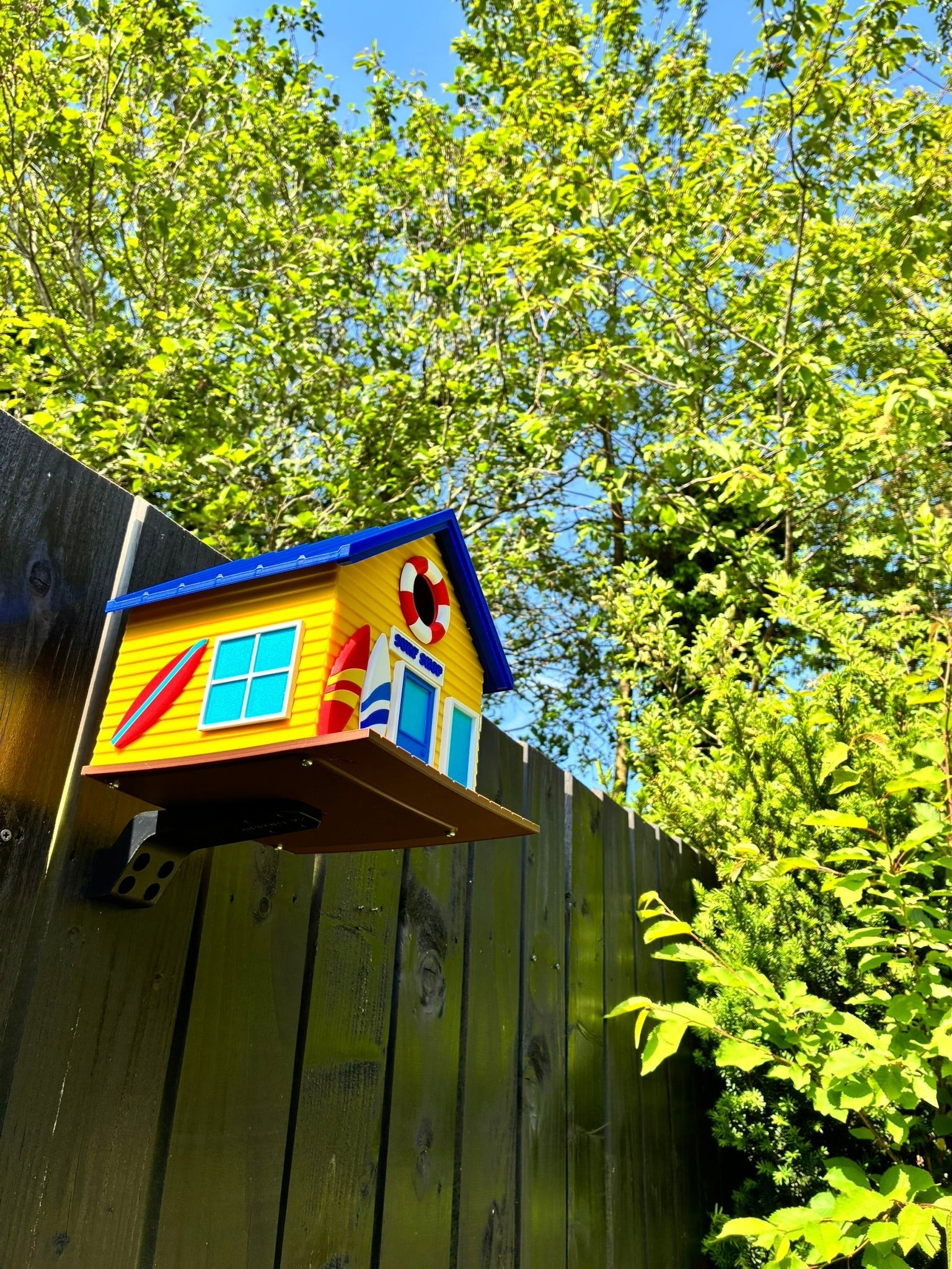 TL3D Prints yellow surf shop birdhouse with blue roof, surfboards, and lifebuoy detail, mounted on a fence.