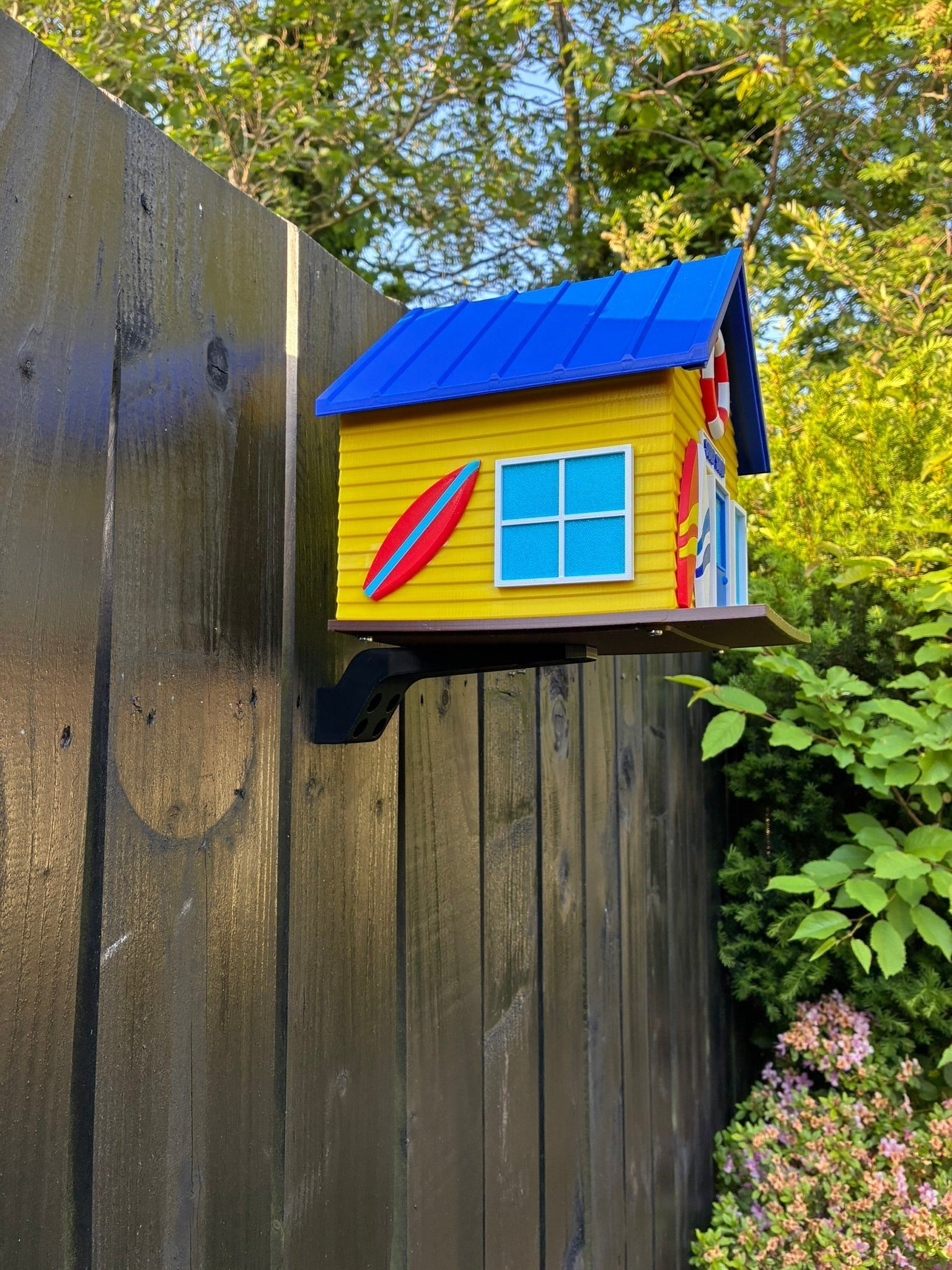 TL3D Prints yellow 3D printed surf shop birdhouse with blue roof, surfboard details, and lifebuoy entrance, mounted on a fence.