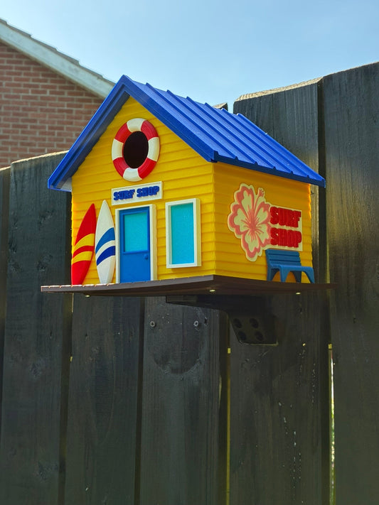TL3D Prints yellow 3D printed surf shop birdhouse with blue roof, surfboards, lifebuoy, and tropical signage.