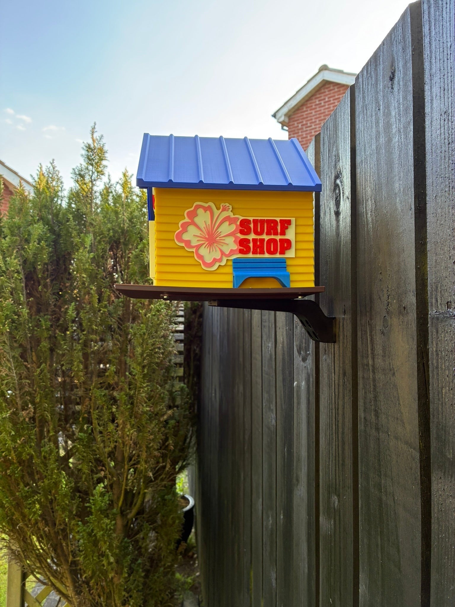 TL3D Prints yellow 3D printed surf shop birdhouse with blue roof, hibiscus flower and 'Surf Shop' sign.