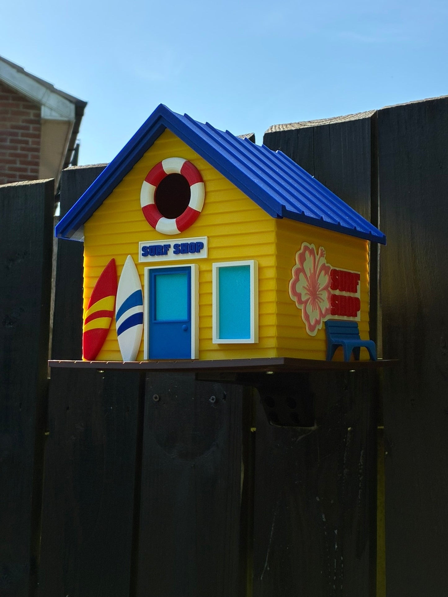TL3D Prints yellow 3D printed surf shop birdhouse with blue roof, surfboards, and lifebuoy detail.