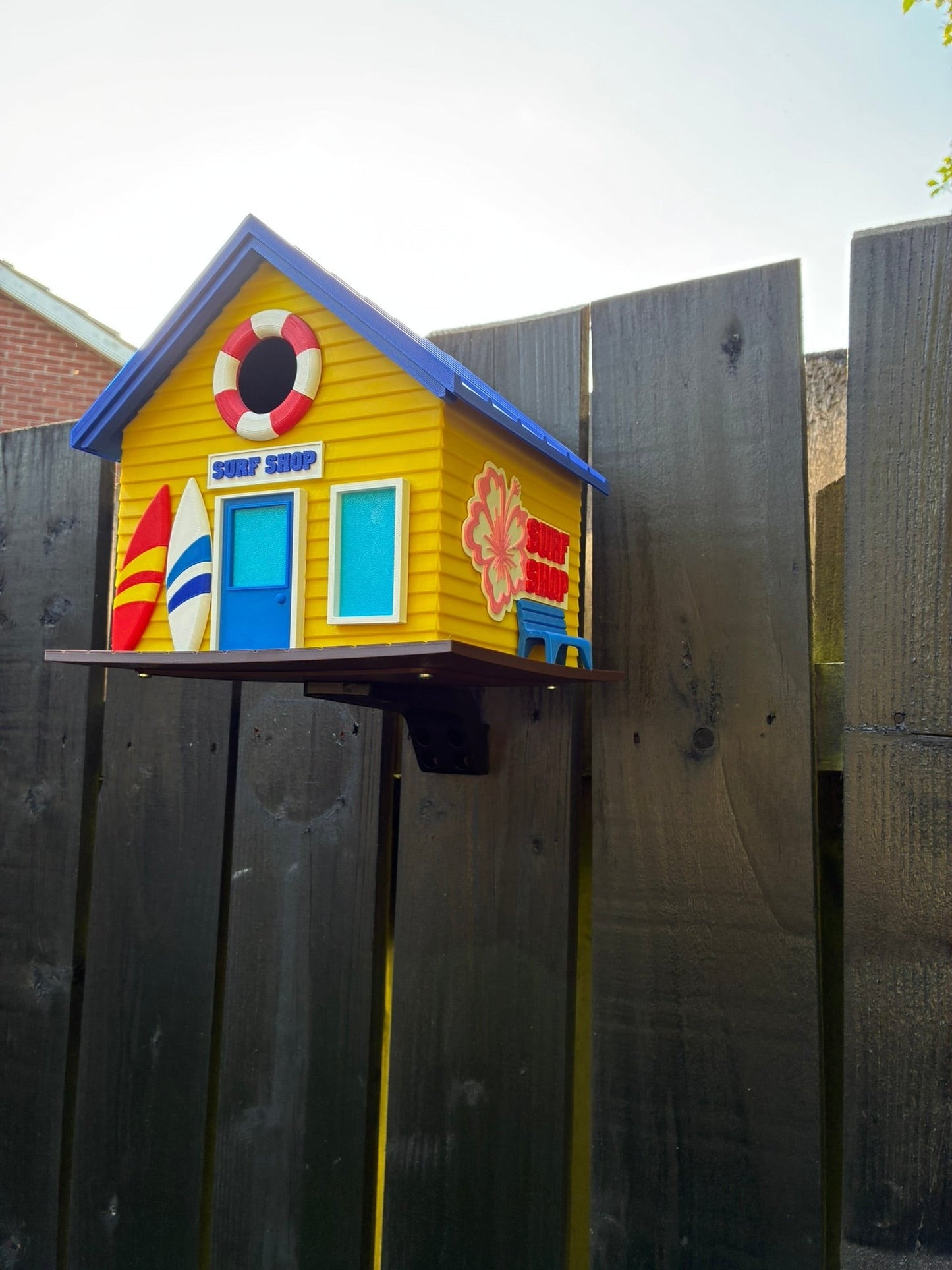 TL3D Prints yellow 3D printed surf shop birdhouse with blue roof, surfboards, and lifebuoy detail. Functional outdoor decor.